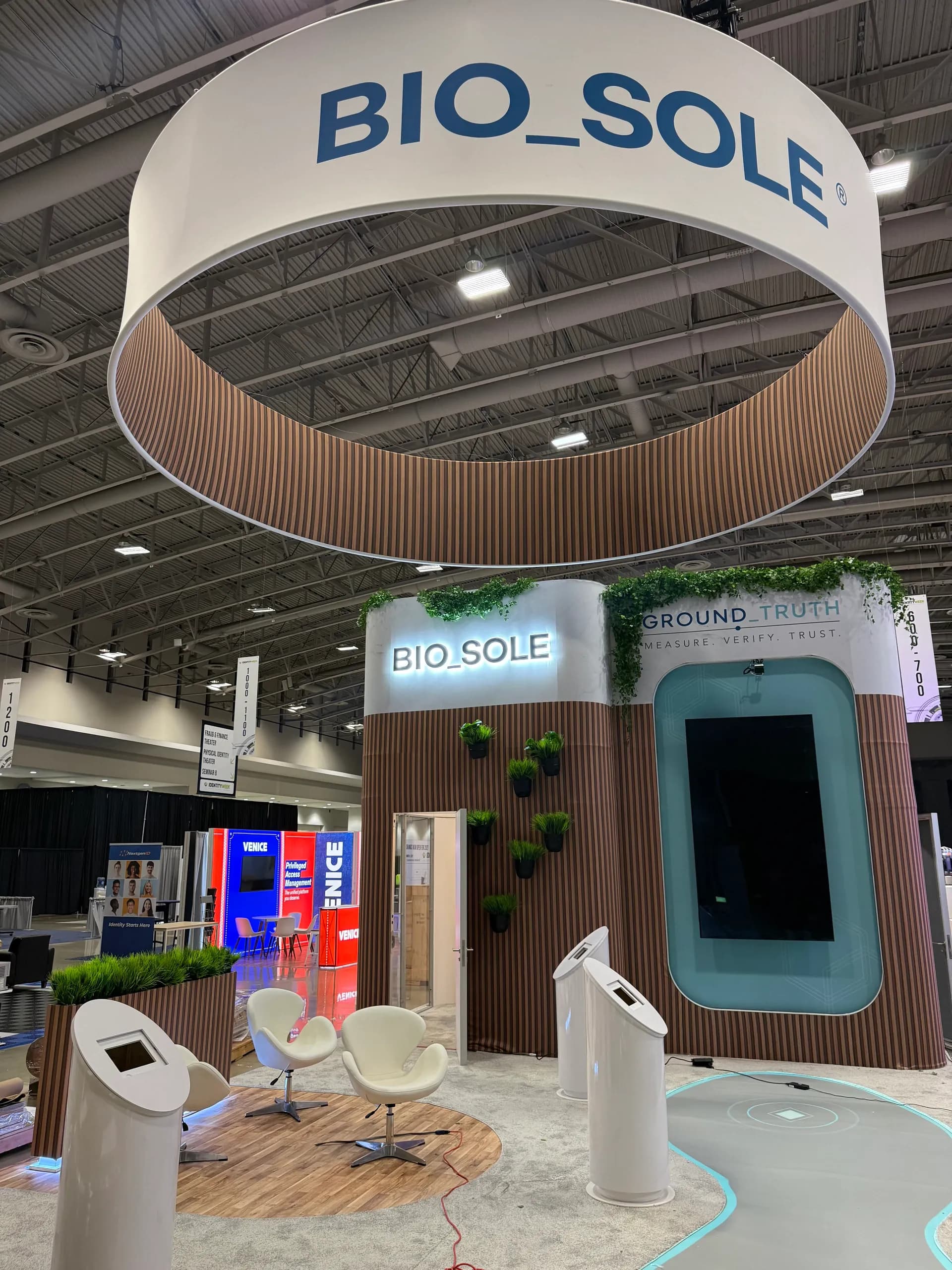 El frente del stand de Bio_Sole: un rótulo BIO_SOLE iluminado y una pantalla Ground_Truth integrados en listones de madera con vegetación, dos sillones blancos y postes de verificación independientes delante